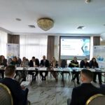 Panel of ten people seated at a long conference table in a meeting room, with a projector screen displaying a presentation behind them.