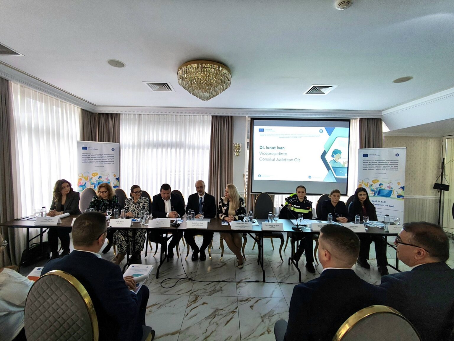 Panel of ten people seated at a long conference table in a meeting room, with a projector screen displaying a presentation behind them.