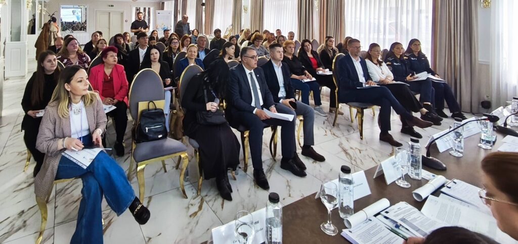 Audience seated in a bright conference room, facing a panel at the front with papers and water bottles visible on the tables.