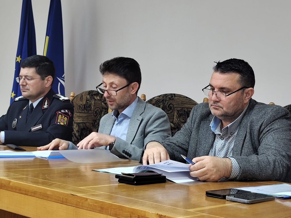Three men sit at a conference table reviewing documents; one in a police uniform, flags visible in the background.