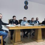 Six men sit at a long wooden table in a meeting room, including a uniformed officer; documents and notebooks are on the table, emblems on the wall behind them.