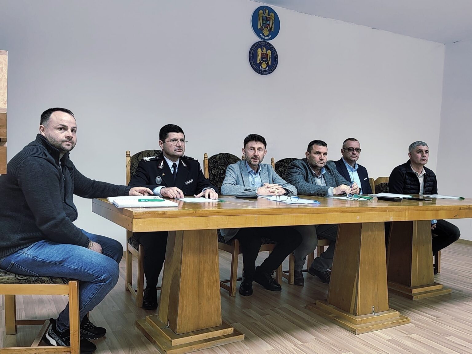 Six men sit at a long wooden table in a meeting room, including a uniformed officer; documents and notebooks are on the table, emblems on the wall behind them.