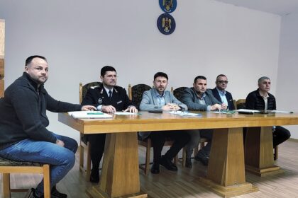 Six men sit at a long wooden table in a meeting room, including a uniformed officer; documents and notebooks are on the table, emblems on the wall behind them.