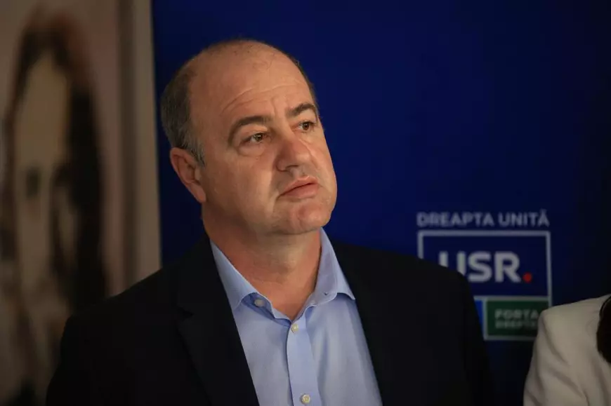 Middle-aged man in a dark blazer and light blue shirt, looking to the side, standing in front of a blue backdrop with a political party logo (USR).