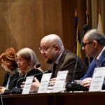 Panel of five professionals at a conference table, speaking into microphones with nameplates in front.