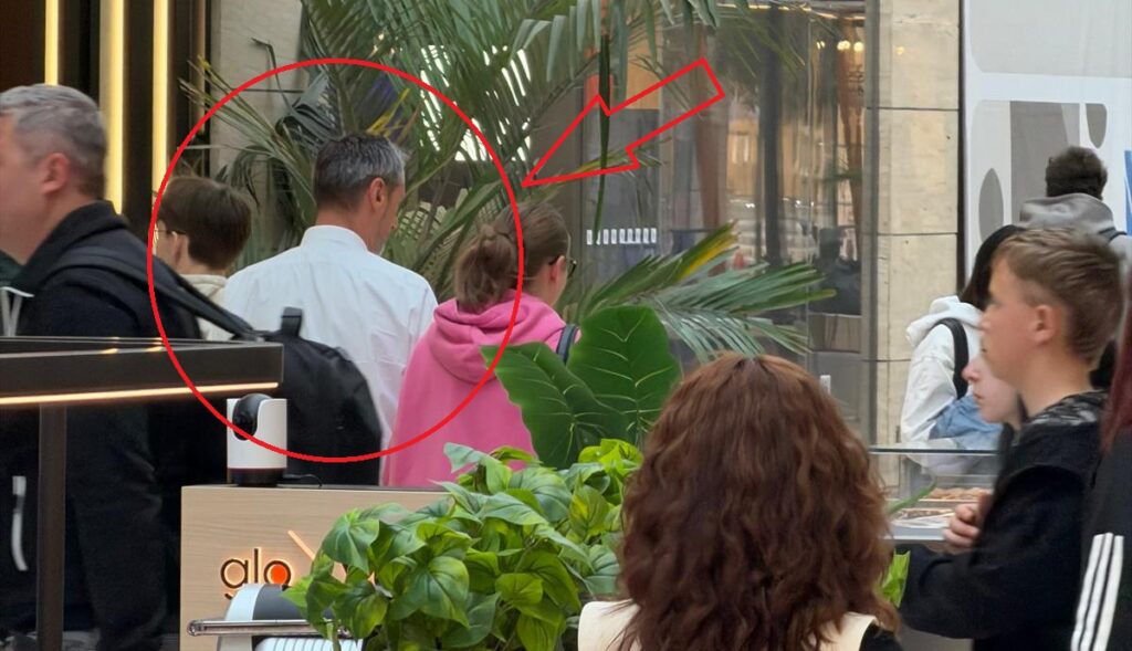 Shoppers in an indoor mall; a man in a white shirt is seen from behind among others, with large tropical plants in the background.