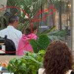 Shoppers in an indoor mall; a man in a white shirt is seen from behind among others, with large tropical plants in the background.