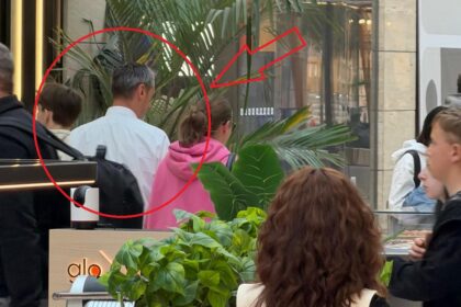 Shoppers in an indoor mall; a man in a white shirt is seen from behind among others, with large tropical plants in the background.