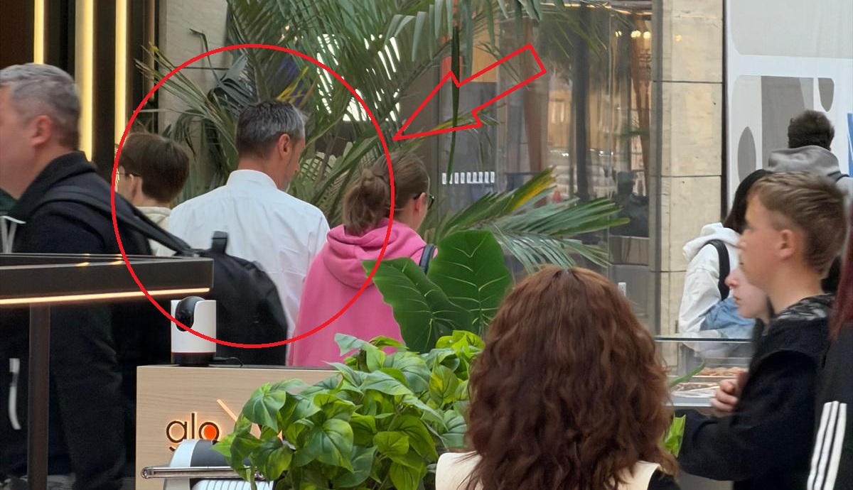 Shoppers in an indoor mall; a man in a white shirt is seen from behind among others, with large tropical plants in the background.