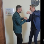 Two men in business attire shake hands and smile in a hallway, as if greeting or celebrating a moment together