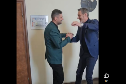 Two men in business attire shake hands and smile in a hallway, as if greeting or celebrating a moment together