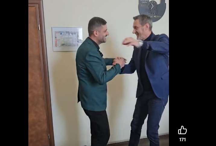 Two men in business attire shake hands and smile in a hallway, as if greeting or celebrating a moment together