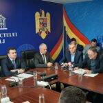 Group of officials in suits seated around a conference table during a government briefing in a blue room with Romanian emblems on the wall.