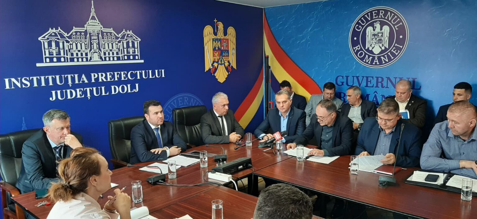 Group of officials in suits seated around a conference table during a government briefing in a blue room with Romanian emblems on the wall.