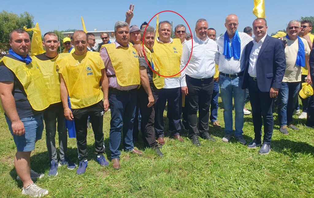 Group of men posing for a photo at an outdoor event; several wear yellow vests with a logo, standing on grass under a sunny sky.