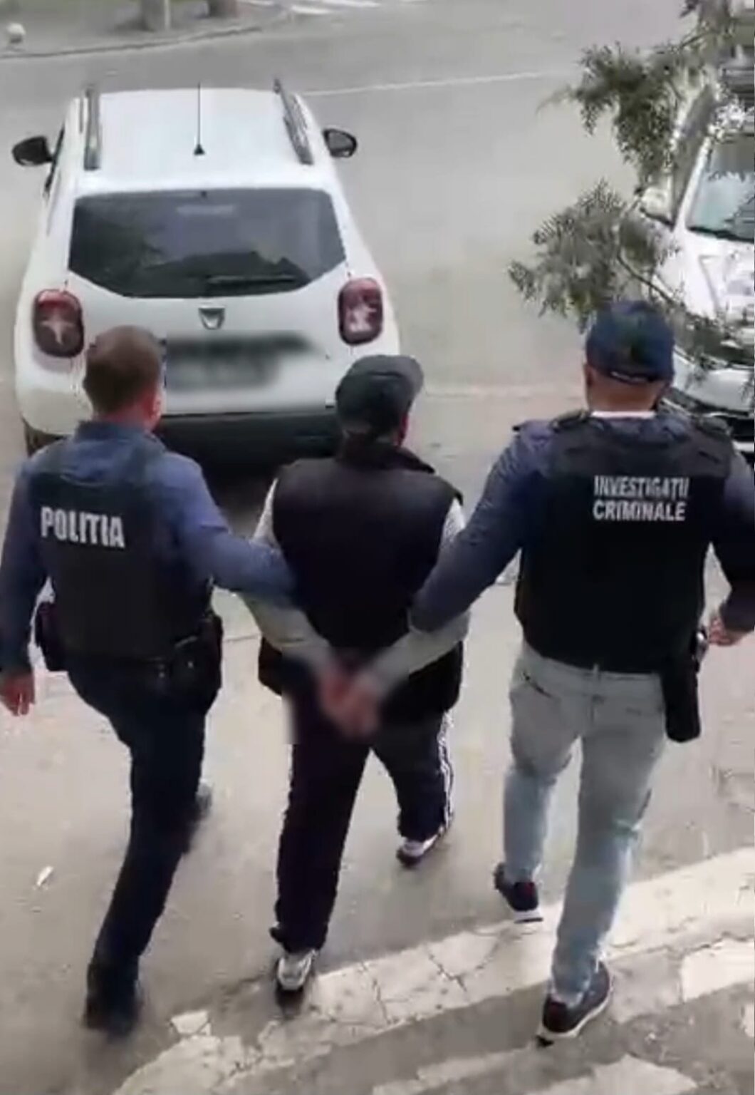 Three officers escort a handcuffed man down a sidewalk toward a white car; vests read 'Politia' and 'Investigatii Criminale' in a street scene.