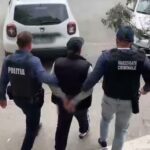 Three officers escort a handcuffed man down a sidewalk toward a white car; vests read 'Politia' and 'Investigatii Criminale' in a street scene.