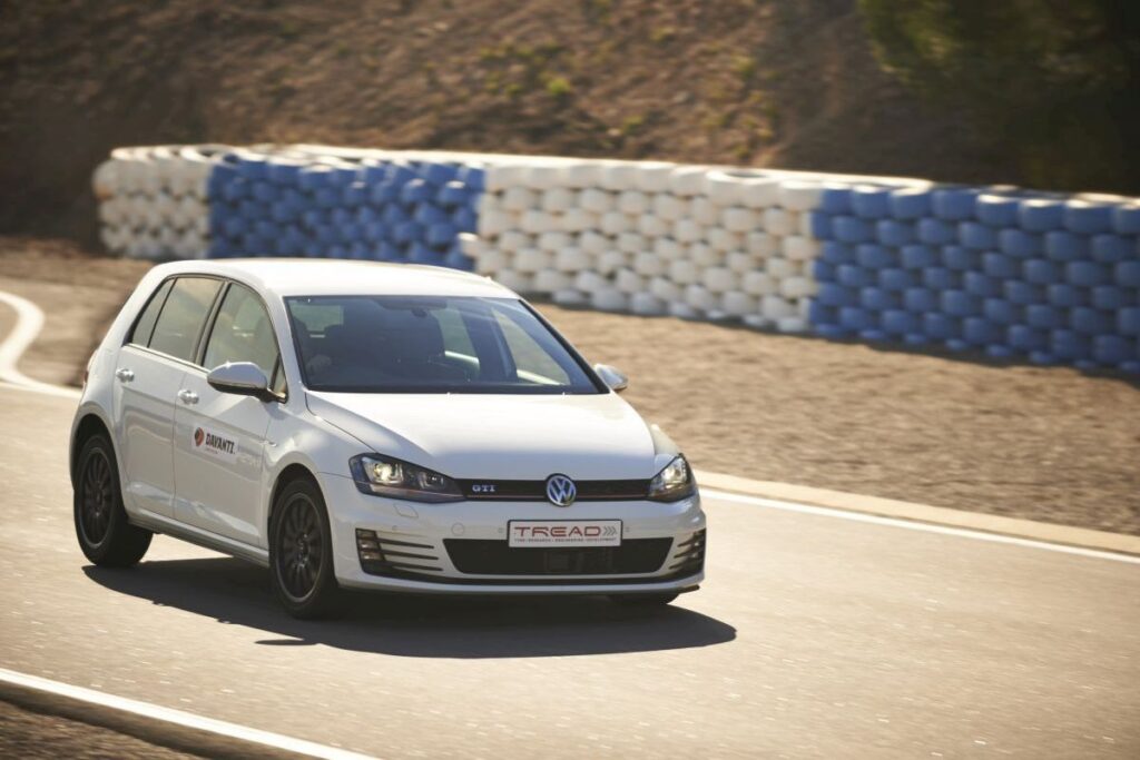 White Volkswagen Golf GTI driving on a racetrack with blue-and-white tire barriers in the background.