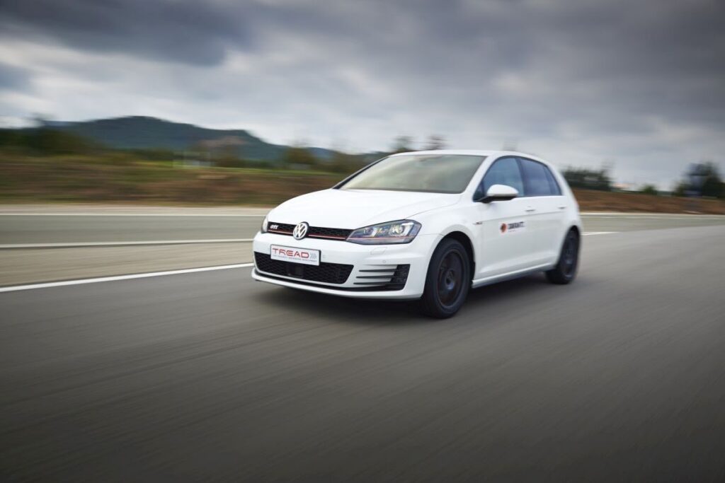 White Volkswagen hatchback driving at speed on a highway under a cloudy sky, motion blur visible.
