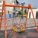 Orange metal swing frame in a park, wrapped in red-and-white caution tape; playground area with buildings in the background.