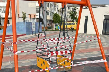 Orange metal swing frame in a park, wrapped in red-and-white caution tape; playground area with buildings in the background.