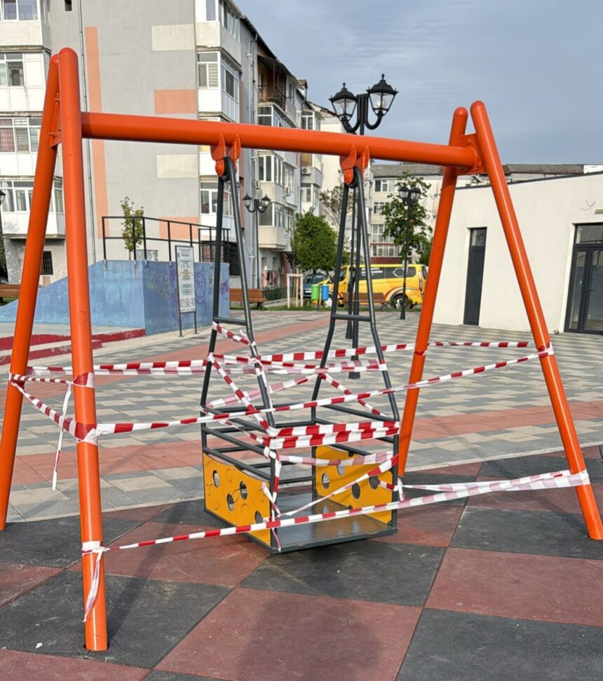 Orange metal swing frame in a park, wrapped in red-and-white caution tape; playground area with buildings in the background.
