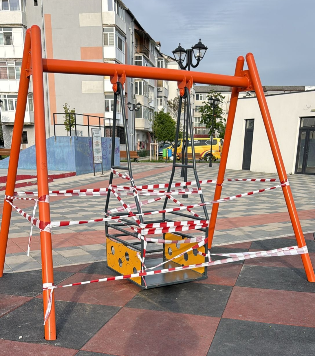 Orange metal swing frame in a park, wrapped in red-and-white caution tape; playground area with buildings in the background.