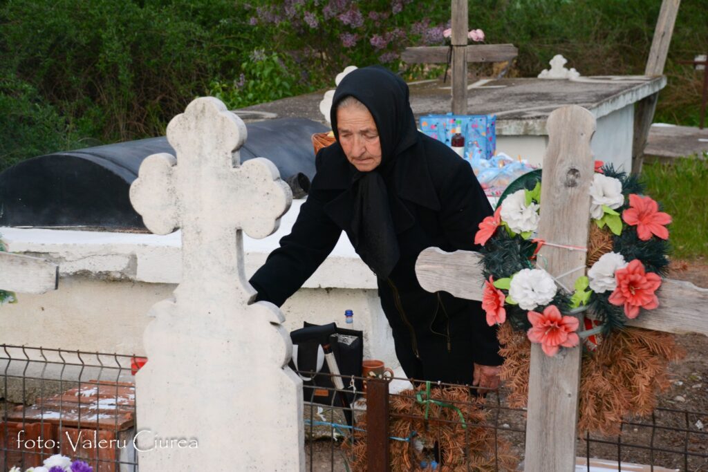Elderly woman in a black headscarf tending a grave with flowers and a cross nearby.
