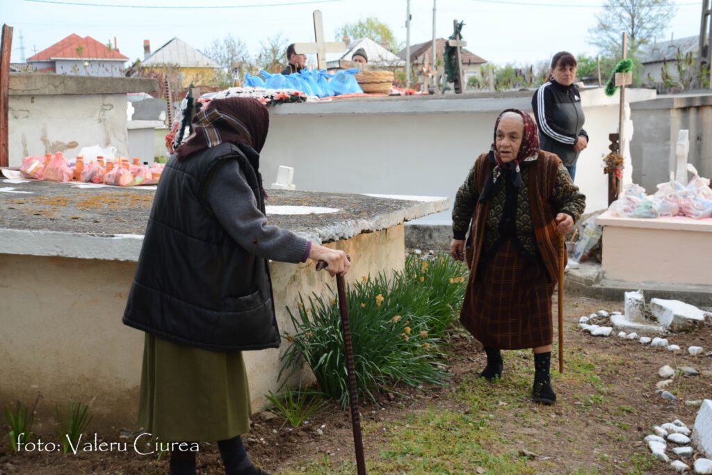 Two elderly women in headscarves stand near graves with crosses and flower offerings in a rural cemetery; one woman leans on a walking staff while another watches from the background.