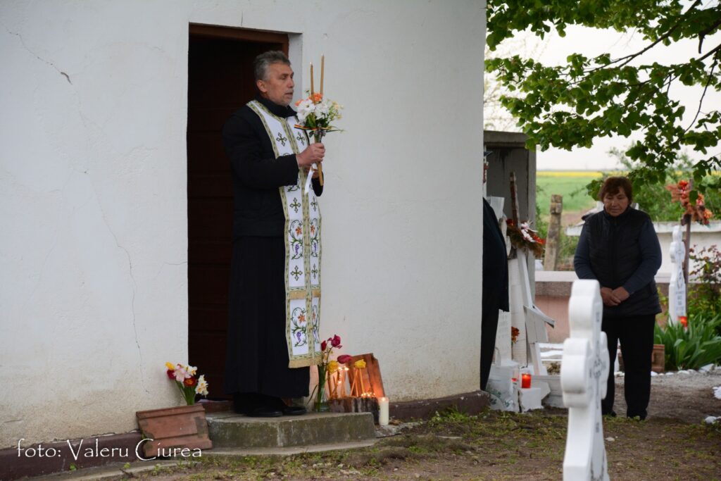 Priest in ceremonial vestments holding candles and flowers at a doorway, with a woman in a vest nearby near a cemetery area.