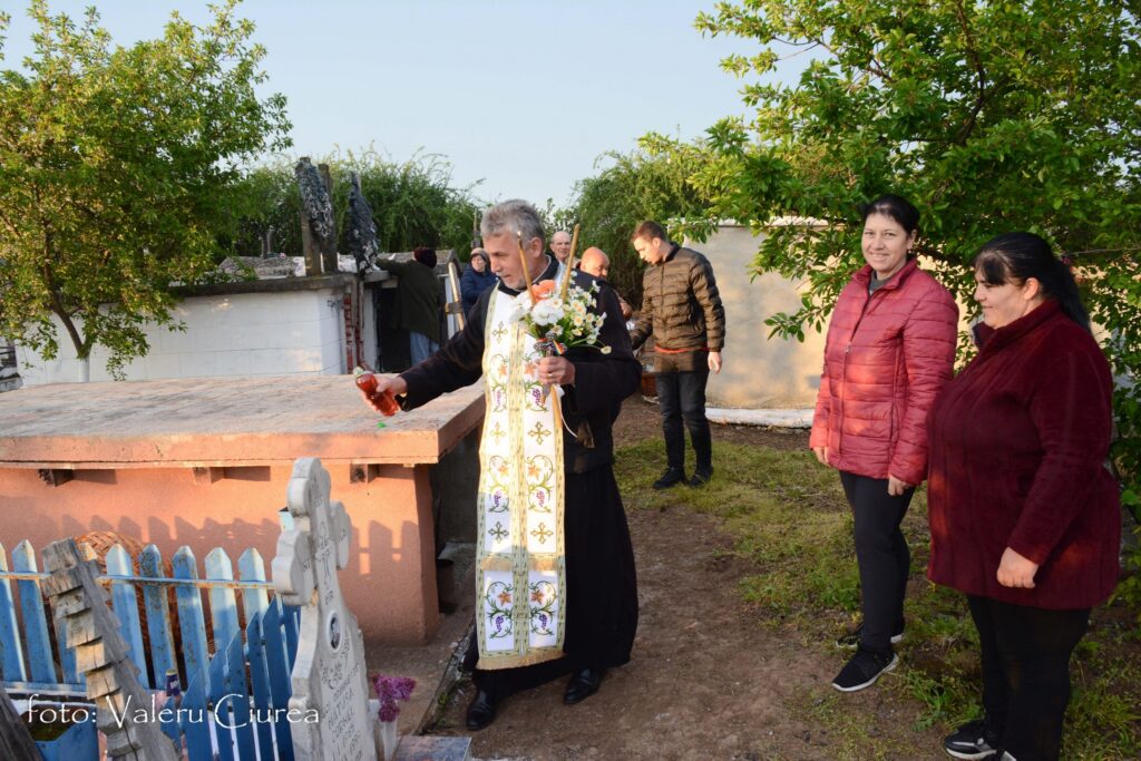 Priest in ornate vestments blesses a grave with flowers while a small group watches nearby in a sunny cemetery.