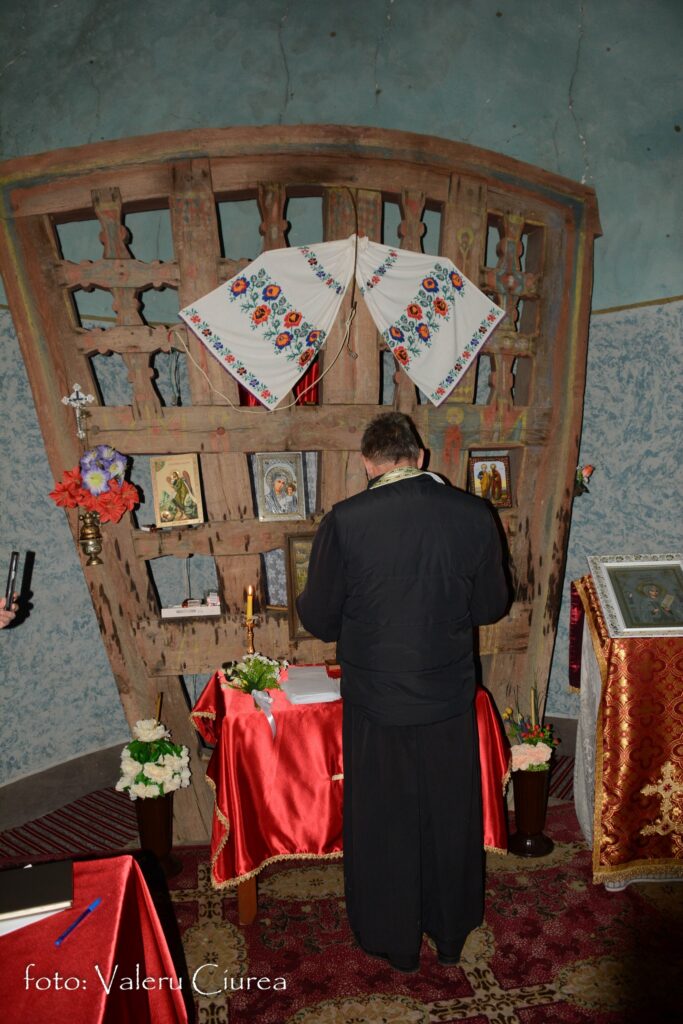 Man in dark robes praying before a carved wooden icon stand, adorned with embroidered cloth and religious icons in a church interior.