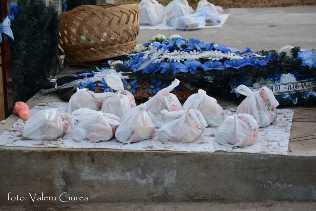 Outdoor memorial display featuring blue artificial flowers and white ribbons on a platform, with a wicker basket and white plastic bags nearby.