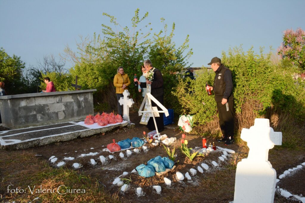 Mourners at a gravesite with white crosses, flowers, and bags of offerings in a sunny outdoor setting