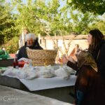 People sit around a table outdoors with bags of groceries and baskets on a sunny day under trees in a yard or garden setting.