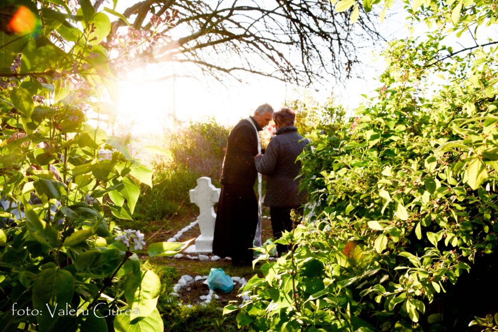 Two people in dark coats stand by white grave markers in a sunlit garden cemetery, framed by green bushes and leaves.