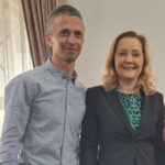 Two adults pose for a photo indoors, smiling at the camera; man in a light blue shirt and the woman in a black blazer with a green patterned blouse.