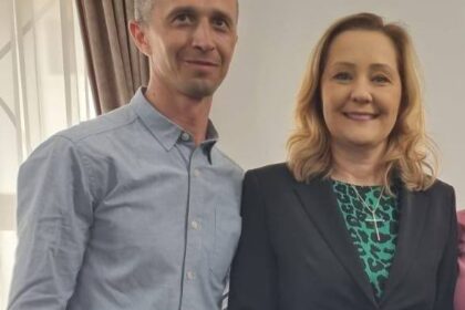Two adults pose for a photo indoors, smiling at the camera; man in a light blue shirt and the woman in a black blazer with a green patterned blouse.