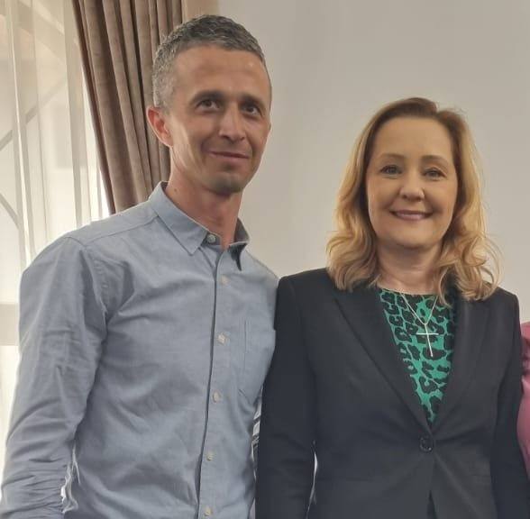 Two adults pose for a photo indoors, smiling at the camera; man in a light blue shirt and the woman in a black blazer with a green patterned blouse.