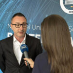 Man in a black suit and white shirt speaks into a blue microphone held by a reporter, in front of a blue interview backdrop with white text.