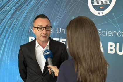 Man in a black suit and white shirt speaks into a blue microphone held by a reporter, in front of a blue interview backdrop with white text.
