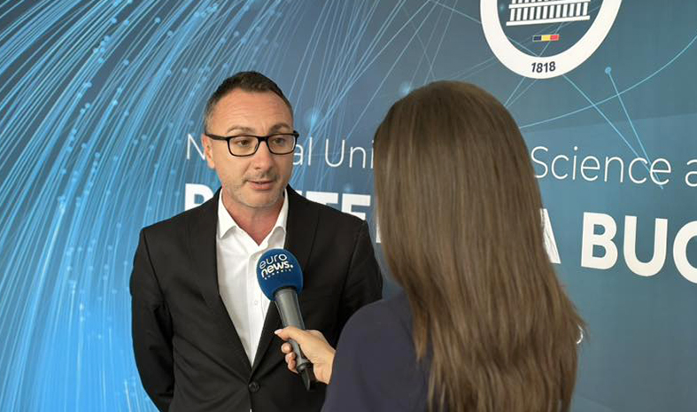 Man in a black suit and white shirt speaks into a blue microphone held by a reporter, in front of a blue interview backdrop with white text.