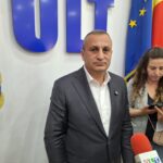 Man in a dark pinstripe suit at a press event, with microphones and reporters nearby, European flags in the background.