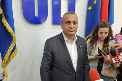 Man in a dark pinstripe suit at a press event, with microphones and reporters nearby, European flags in the background.