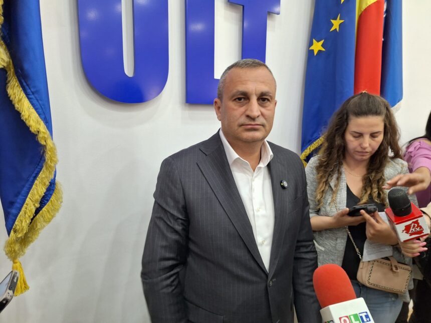 Man in a dark pinstripe suit at a press event, with microphones and reporters nearby, European flags in the background.