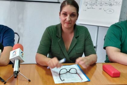 A woman in a green blazer sits at a wooden table during a press briefing, with a microphone labeled TVR, papers, and reading glasses on the table.