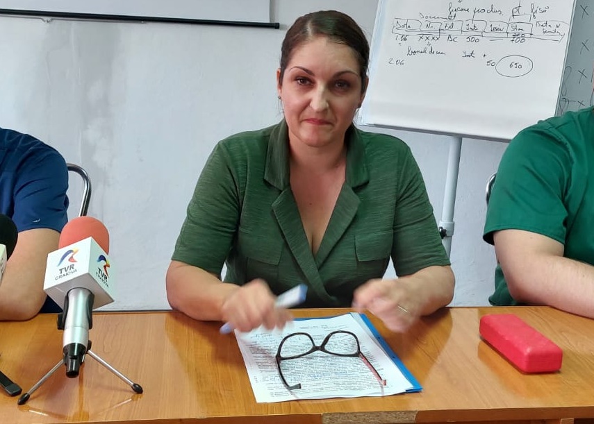 A woman in a green blazer sits at a wooden table during a press briefing, with a microphone labeled TVR, papers, and reading glasses on the table.