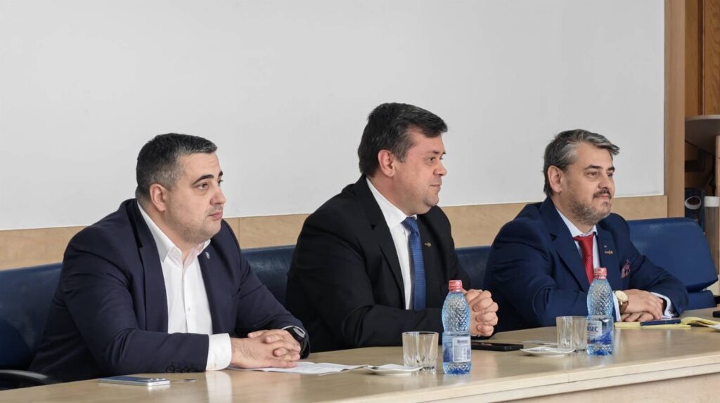 Three men in suits seated at a conference table during a meeting, with water bottles and papers on the table in front of them.