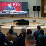 Large screen showing a man in a suit delivering a speech, with smaller thumbnails of other attendees below, in a conference hall with a circular seating arrangement.