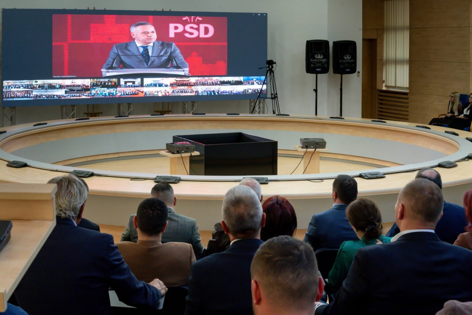 Large screen showing a man in a suit delivering a speech, with smaller thumbnails of other attendees below, in a conference hall with a circular seating arrangement.
